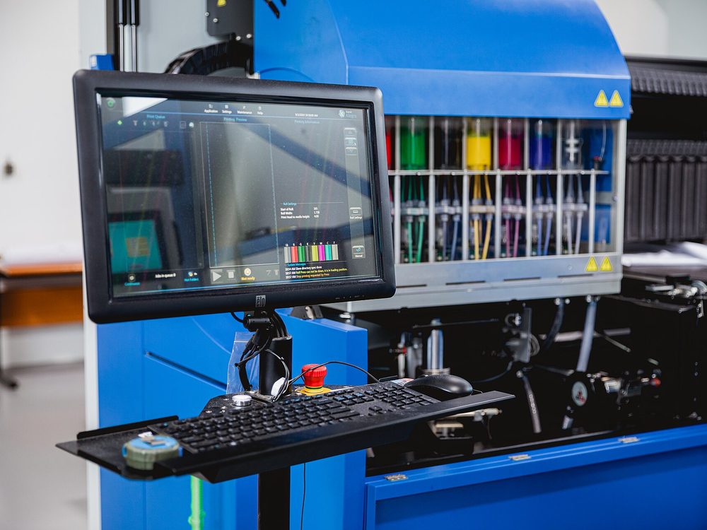 Control screen of a digital textile printer showing the print queue, with the ink supply behind it