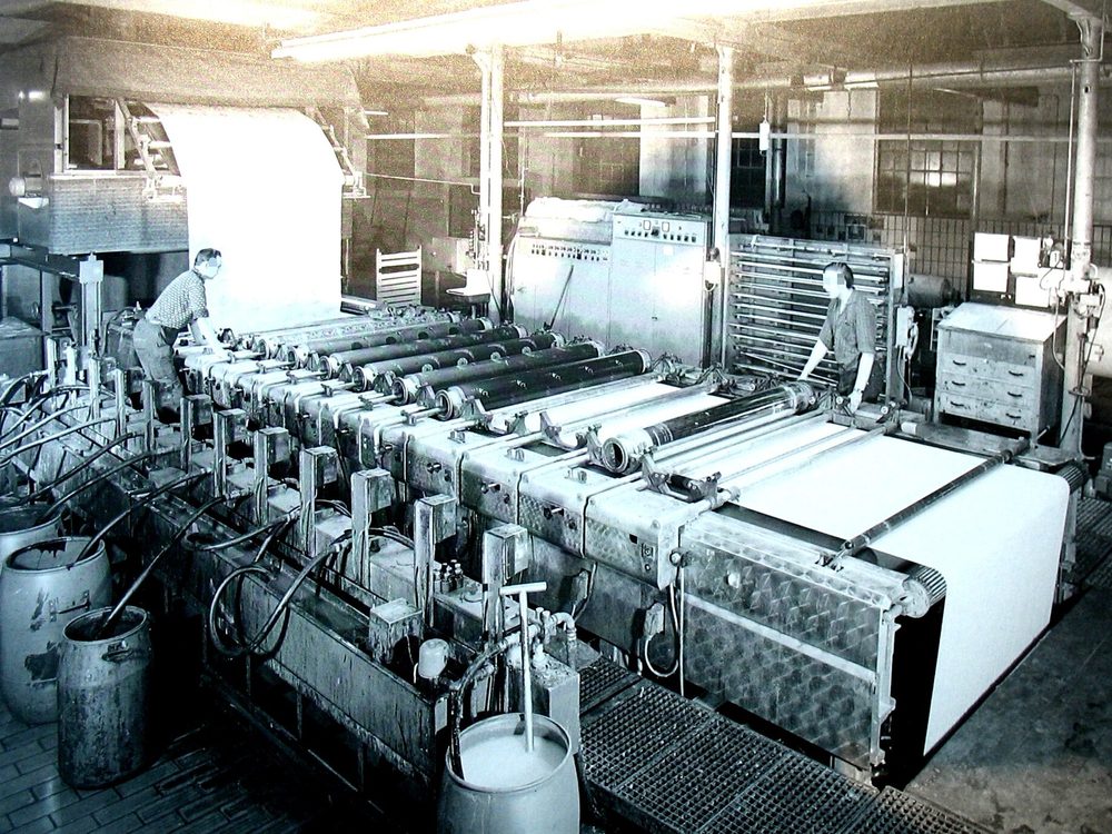 Black and white photograph of a rotary screen printing line with a row of cylindrical screens over white cloth